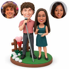 custom bobblehead figures singing couple with a man holding a microphone on a stand beside a woman gesturing in front of a heart-topped wooden fence