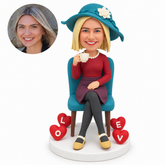 custom bobblehead figure woman seated with crossed legs drinking tea beside four lettered hearts