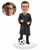 custom bobblehead figure male judge holding a gavel across his waist with both hands and resting his right foot on a soccer ball