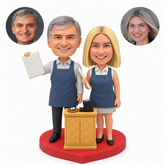 Custom couple bobblehead in service aprons beside a cash counter, with the man raising an order board