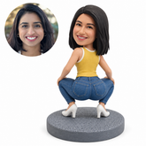 Custom soft-clay bobblehead woman in a deep rear-facing squat, wearing a muted yellow sleeveless top, blue jeans and white heels on a gray round base, with matching real-photo portrait.