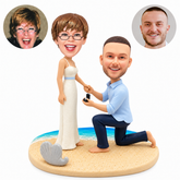 Custom clay beach proposal couple: woman standing in ivory dress, man kneeling with open ring box, sandy shoreline base and seashell plaque, with matching portrait photos
