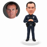 Custom clay police officer bobblehead standing with both empty hands clasped high at the chest in a charcoal-navy uniform, with matching real portrait example.
