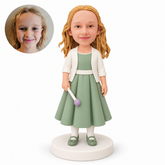 Custom polymer-clay bobblehead girl in a sage green pleated gown holding one lowered round-tipped wand