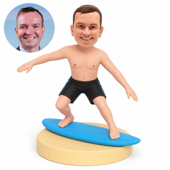 Custom clay bobblehead of a shirtless man balancing on a blue surfboard over a sand base