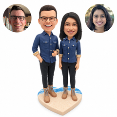 Custom bobblehead couple in matching slate-blue shirts and lace-up boots on a heart-shaped beach base