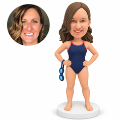 Custom polymer-clay woman swimmer in a navy swimsuit with both hands on hips and one pair of goggles hanging by its strap from her right wrist, with matching portrait photo