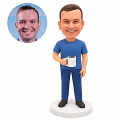 Custom polymer-clay man in a blue T-shirt holding a white mug by its handle in his right hand and a slim grey rectangular accessory in his lowered left hand, on a plain round base, with matching real portrait