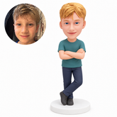 Custom polymer-clay boy in a teal T-shirt and navy trousers, standing with folded arms and his left leg crossed in front on its forefoot, on a plain round base, with matching real child portrait