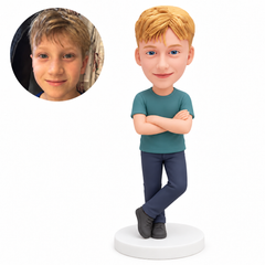 Custom polymer-clay boy in a teal T-shirt and navy trousers, standing with folded arms and his left leg crossed in front on its forefoot, on a plain round base, with matching real child portrait