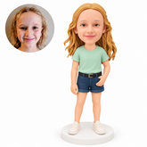 Custom polymer-clay girl in a mint T-shirt and navy shorts with her left palm exposed across the front pocket and her right empty arm down, on a plain round base, with matching real child portrait