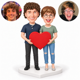 Two clay figurines share one large red heart with lower side grips, standing on a white heart-shaped base with matching male and female real portraits.