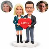 Couple clay figures hold a red LOVE heart with two side grips and one hand on top, on a white heart-shaped base with matching real portraits.