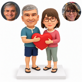 Casual clay couple in sandals sharing a red heart with open front palms and lower supporting hands on a white rectangular base, with matched real portraits.