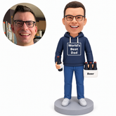 Clay dad in a navy World’s Best Dad hoodie, both arms lowered with one bottle low at his right thigh and a white bottle carrier at his left, gray base and matching real portrait.