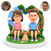 custom bobblehead figures couple seated on a seesaw in front of a heart-shaped sky and tree backdrop