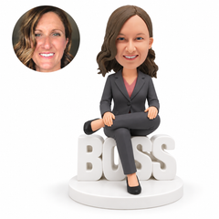 custom bobblehead figure businesswoman seated on BOSS letters with one ankle resting across the opposite knee and both hands apart on her raised leg