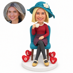 custom bobblehead figure woman seated with crossed legs drinking tea beside four lettered hearts