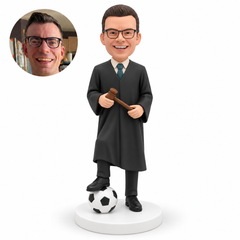 custom bobblehead figure male judge holding a gavel across his waist with both hands and resting his right foot on a soccer ball