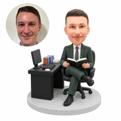 custom bobblehead figure businessman seated cross-legged reading an open book beside a computer desk