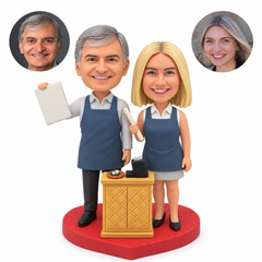 Custom couple bobblehead in service aprons beside a cash counter, with the man raising an order board