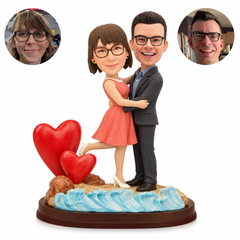 Custom clay couple embracing by two red hearts and sculpted ocean waves, with woman lifting one leg and matching real photo portraits.