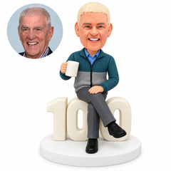 Custom clay bobblehead of an older man seated on 100 birthday numerals with crossed legs and a mug, beside his matching portrait photo.