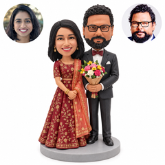 Custom Indian wedding couple clay bobbleheads: bride in berry lehenga clasping her own hands, groom in charcoal suit holding one flower bouquet with both hands, with matching real portrait circles on a round base