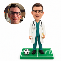 Custom male doctor bobblehead in teal scrubs holding a small ball and book on a soccer field base, with matching round portrait reference.