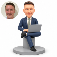 custom bobblehead figure businessman seated on a cylindrical stool with an ankle on his opposite knee using a laptop