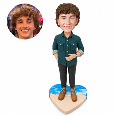Custom clay bobblehead man in a teal shirt with one hand in his pocket and the other across his abdomen on a heart-shaped beach base