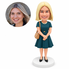 Custom polymer-clay bobblehead woman in a teal dress with empty hands folded in front and a tan right shoulder bag, with matching portrait photo