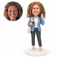 Custom polymer-clay bobblehead woman in a blue blazer holding a takeaway cup in her right hand, raising her empty left hand beside her head and carrying a right shoulder bag, with matching portrait photo