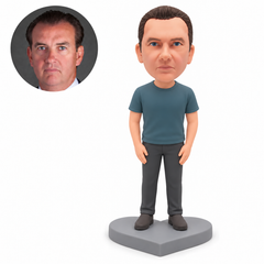 Custom polymer-clay man in a muted teal T-shirt and charcoal trousers with both empty hands lowered beside his thighs, standing on a plain grey heart-shaped base, with his matching real portrait